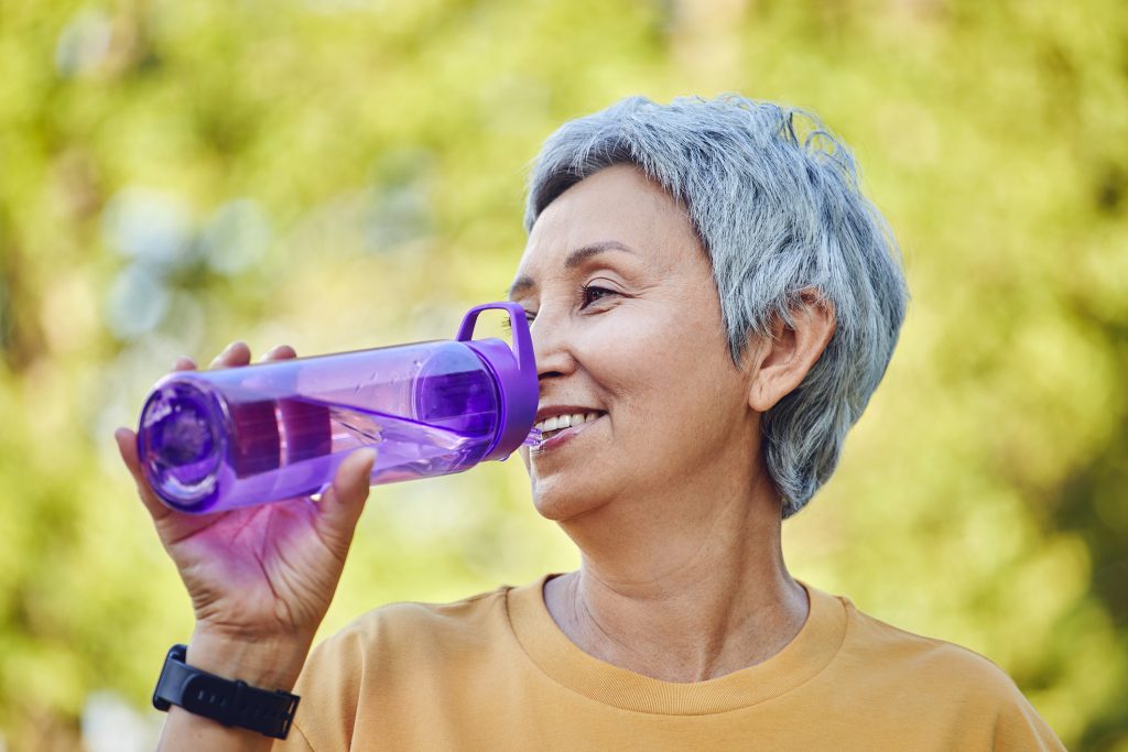 Une femme aux cheveux gris d'origine sud-asiatique affichant un air heureux boit l'eau d'une bouteille mauve