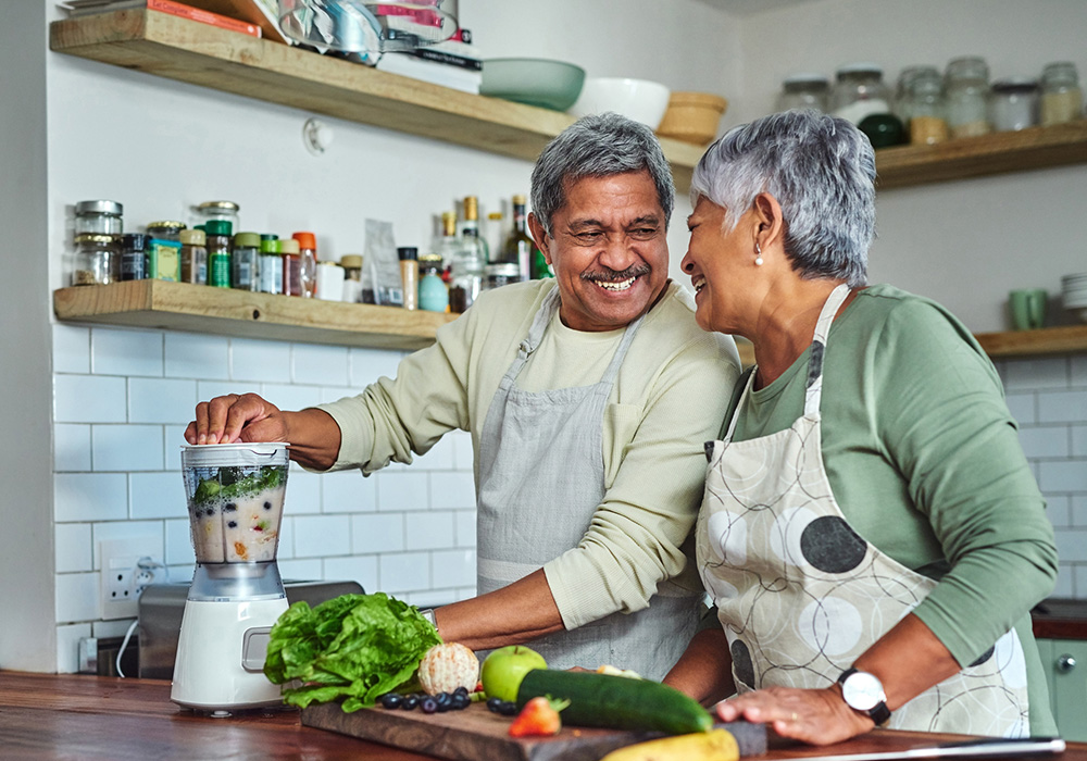 An older couple smiling at each other, preparing a smoothie in their kitchen
