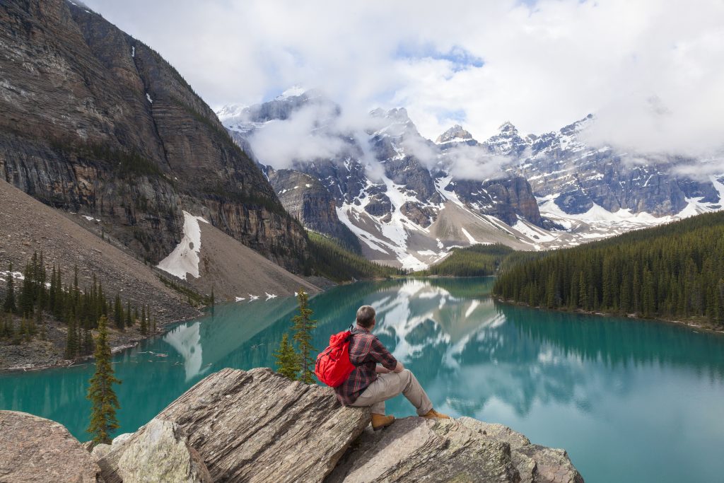 Prise de vue arrière d'un randonneur assis sur des rochers qui regarde les montagnes de l'autre côté de l'étendue d'eau.