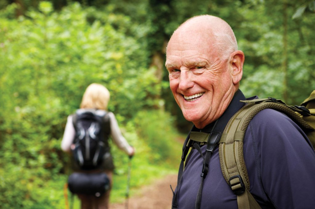 Older man and woman walking in the forest, man in the forefront smiling