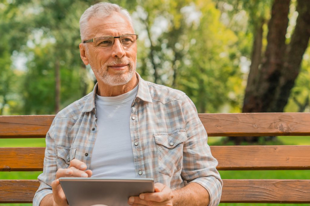 Homme aux cheveux gris assis sur un banc de parc regardant au loin avec une tablette électronique à la main