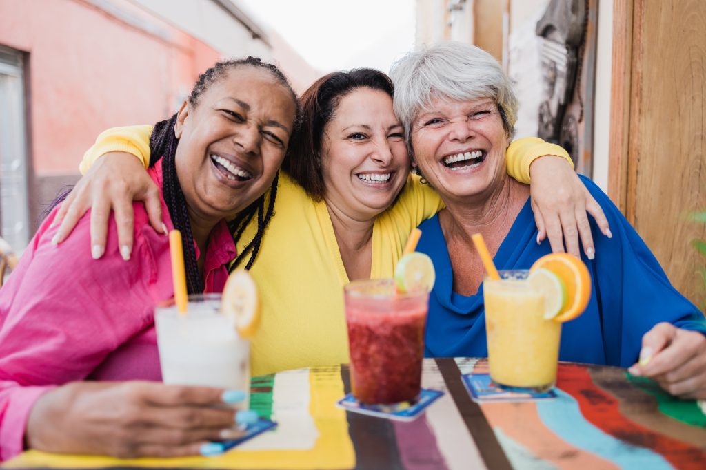 Three older women embracing and laughing with smoothies at restaurant outside