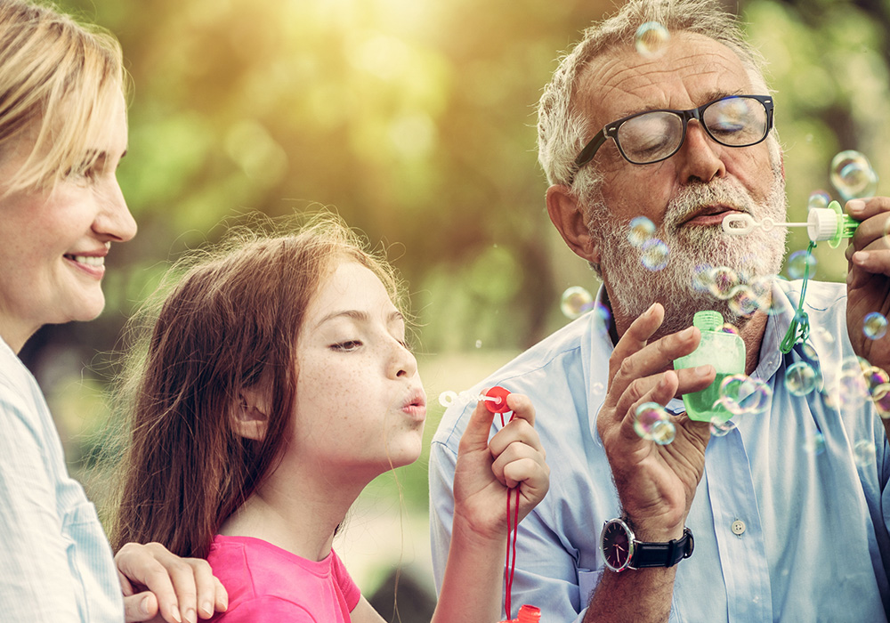 Woman smiling watching older man and granddaugher blow bubbles in the park