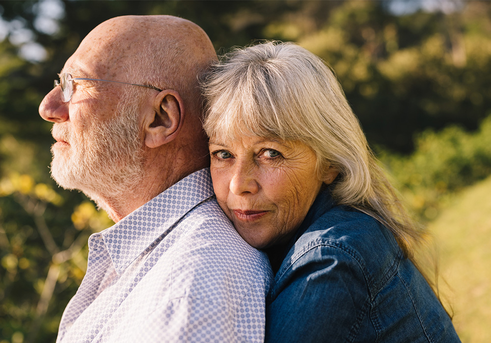 An older woman embraces her husband from behind, staring at the camera.