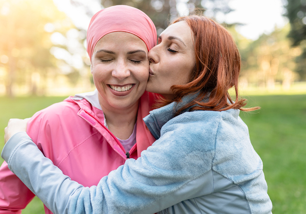 Une femme embrasse affectueusement la joue d'une femme atteinte d'un cancer tout en la serrant dans ses bras à l'extérieur.