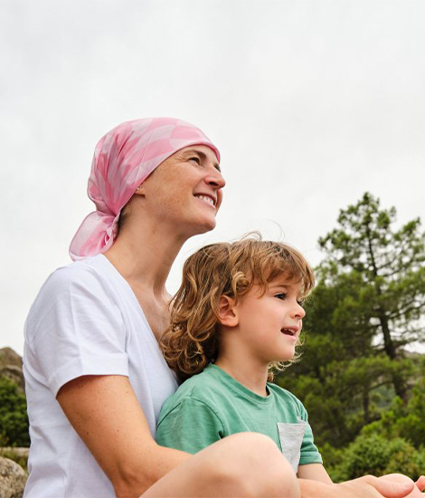 Une jeune femme atteinte d'un cancer est assise à l'extérieur avec un jeune enfant sur ses genoux, elle regarde au loin en souriant.
