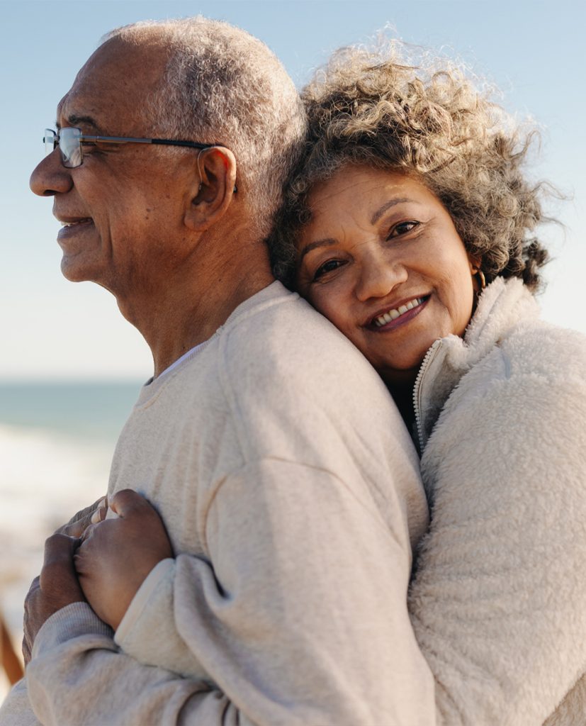 Une femme noire regarde la caméra en souriant en enlaçant son mari devant l'océan.