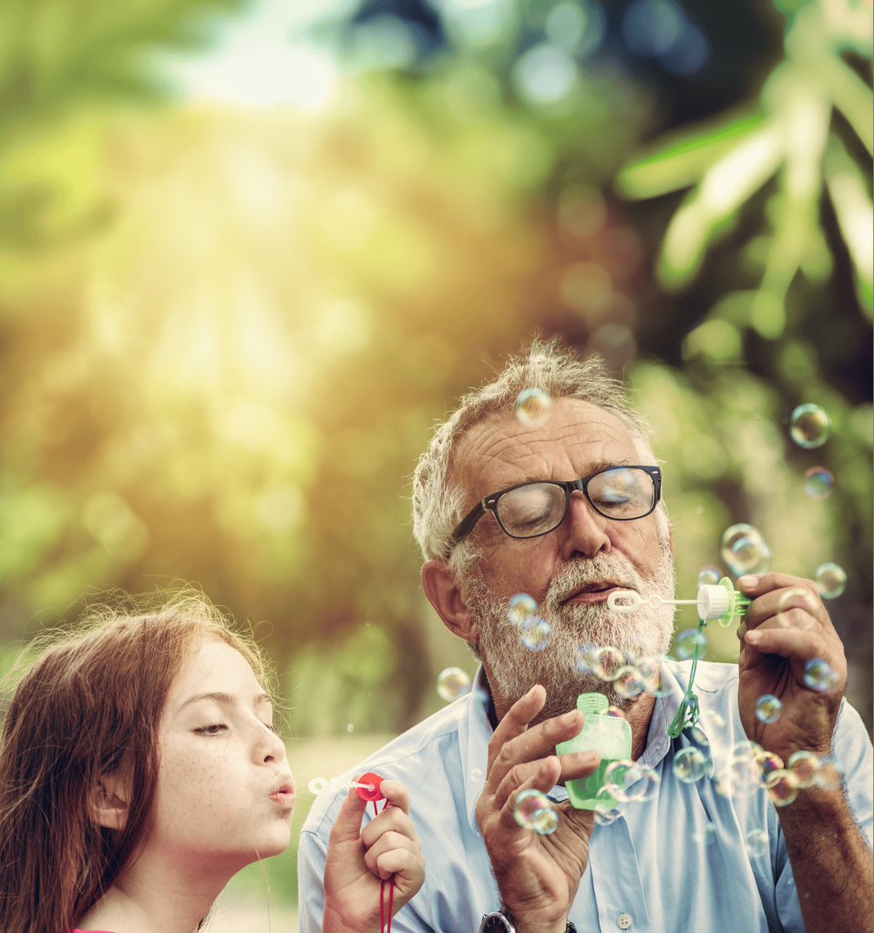 A grandmother smiles as she watches her husband blow bubbles in the park with their granddaughter