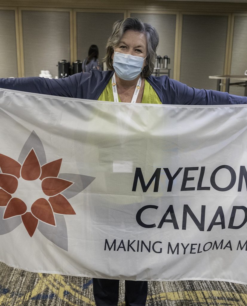 A female Myeloma Canada volunteer wearing a surgical mask during COVID-19 holds a Myeloma Canada banner.