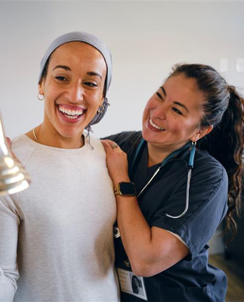 A happy young ethnic girl rings a brass bell to celebrate her last chemotherapy treatment while a nurse proudly holds her shoulder and smiles